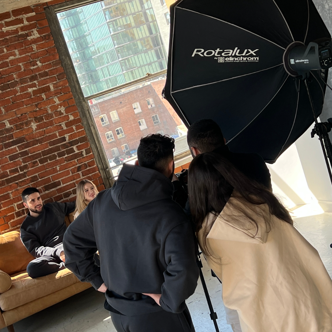 Behind-the-scenes image of a photoshoot for Angels In March streetwear brand, featuring a male and female model sitting on a couch against a brick wall with a window, while a male photographer captures the moment.